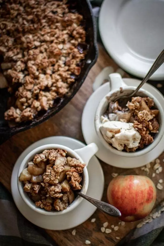 two bowls with apple crisp and vanilla ice cream. In the background is a cast iron skillet with apple crips