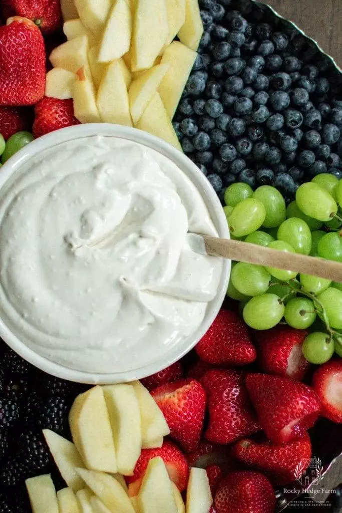 A beautifully arranged fruit platter featuring slices of strawberries, blueberries, blackberries and apples served with healthy cream cheese dip in a small bowl