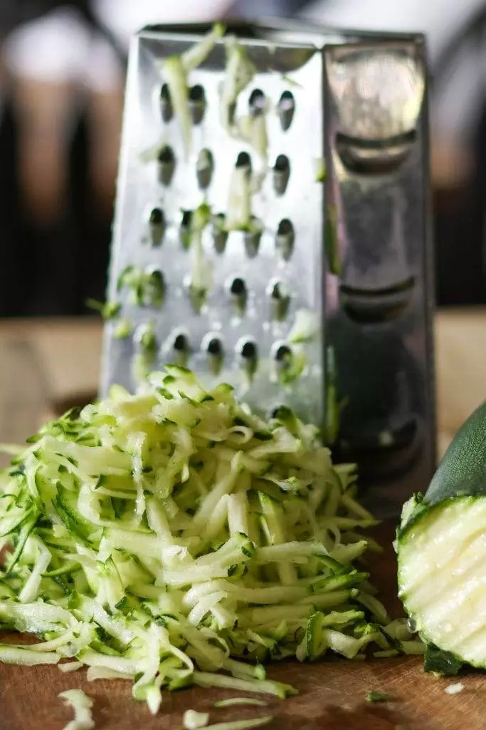 A box grater in the background with a pile of shredded zucchini laying on a cutting board