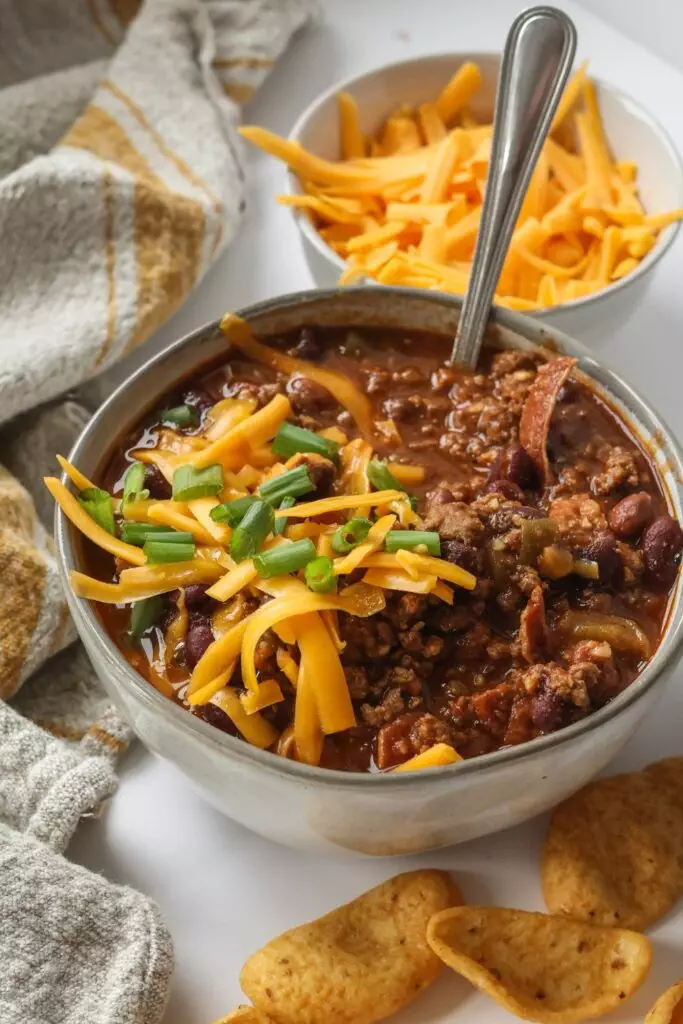 Bowl of delicious chili topped with sour cream, shredded cheddar cheese, and green onions, served with a side of tortilla chips.